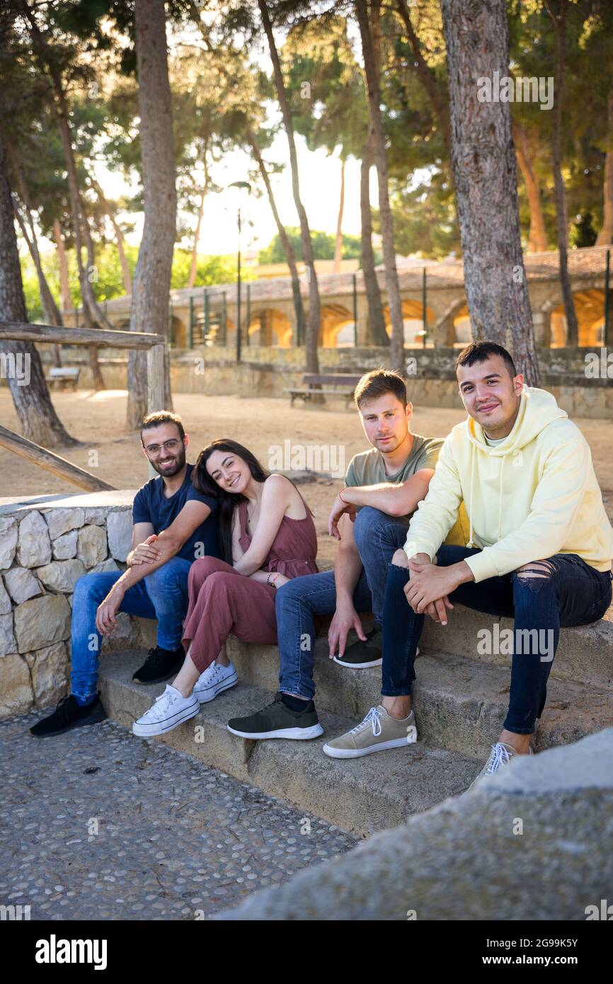 Glückliche Freunde sitzen auf den Stufen eines Parks mit Kiefern. Sie schauen lächelnd auf die Kamera. Sonnenuntergang. Kaukasische, junge Menschen in Sommerkleidung. Jave Stockfoto