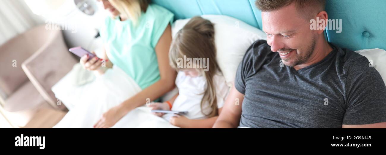 Eltern und Kind liegen im Bett, jeder hält das Smartphone in den Händen Stockfoto