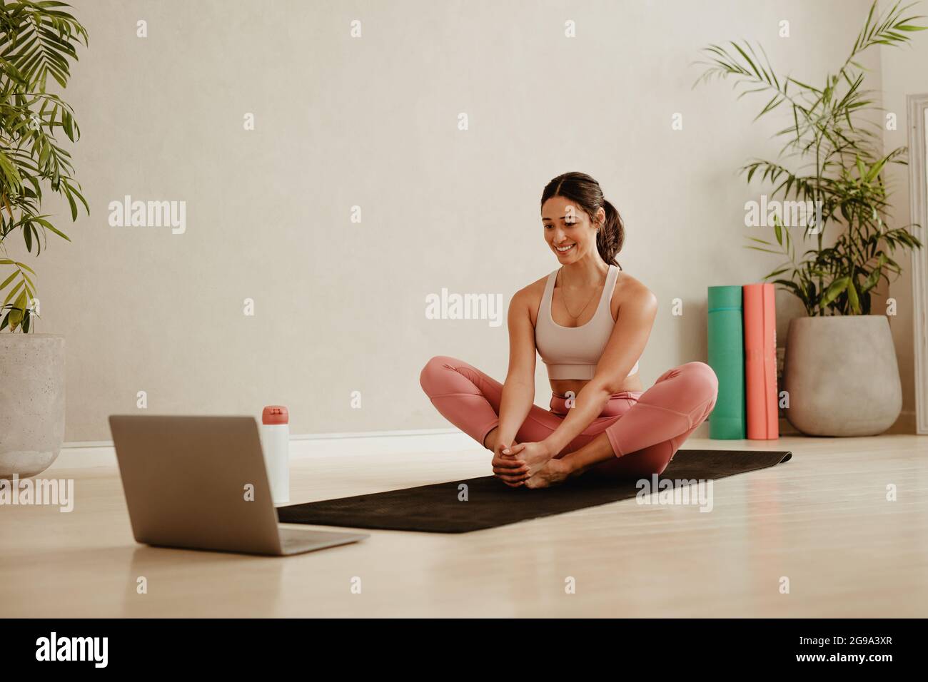 Frau sitzt auf Yoga-Mann und macht Stretching-Training, während Sie Online-Video auf dem Laptop zu Fuß. Sportliche Frau macht zu Hause Übungen, während sie onlin beobachtet Stockfoto