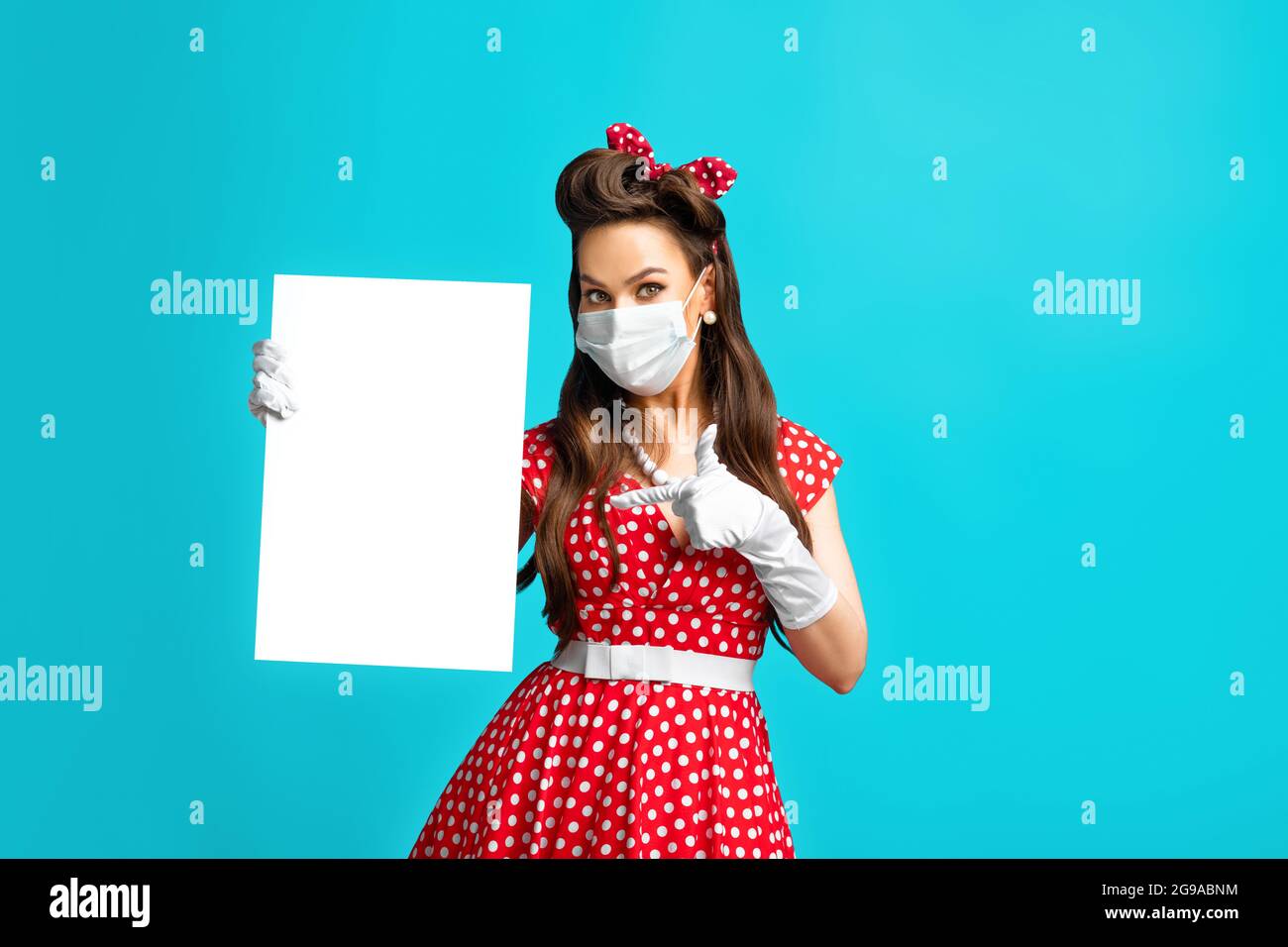 Junge Pinup Frau in Anti covid Gesichtsmaske zeigt auf leeres Papier Poster mit Mockup für Ihr Design auf blauem Hintergrund Stockfoto