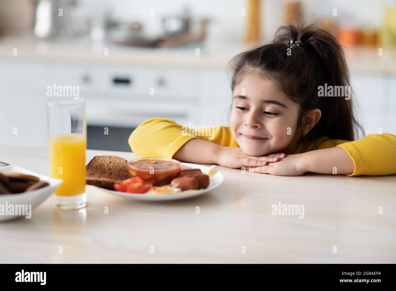 Hungriges Kleines Mädchen, Das Auf Dem Tisch Schief Und Teller Mit Leckerem Essen Ansah Stockfoto