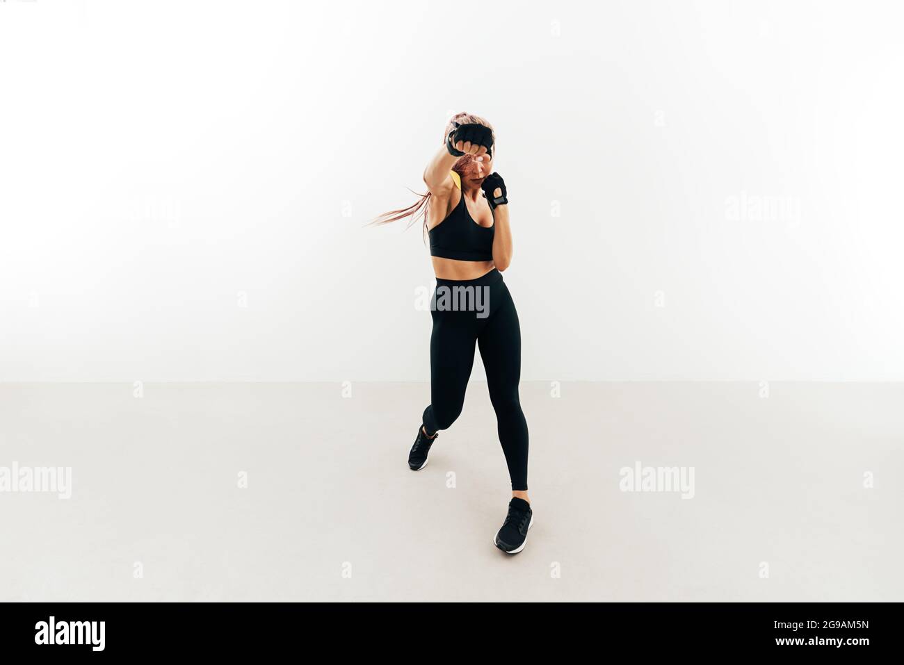 Junge Frau beim Schattenboxen in Innenräumen. Muskulöse Frau in Handschuhen, die gegen eine weiße Wand trainiert. Stockfoto