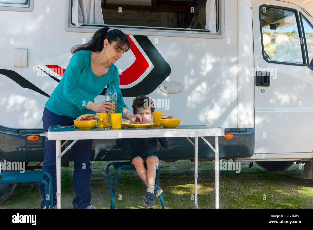 Kind sitzt am Tisch neben dem Wohnmobil, während seine Mutter an einem Campingtag Snacks und Getränke serviert. Stockfoto