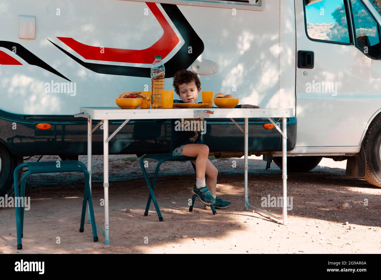 Kleiner Junge im Schatten eines Baumes vor seinem Camper, der auf einem Campingstuhl sitzt und bereit ist, das Essen auf dem Tisch zu essen. Stockfoto