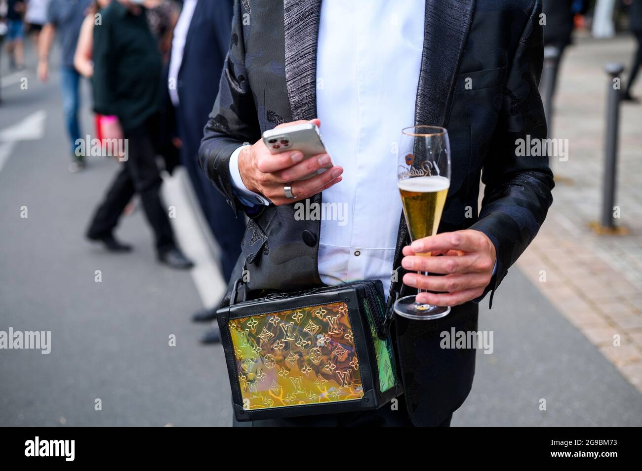 Bayreuth, Deutschland. Juli 2021. Eröffnung der Richard Wagner Festspiele 2021 mit einer Neuproduktion der Oper 'der fliegende Holländer'. Ein Operngast trägt eine schillernde Louis Vuitton Tasche und Blicke auf sein Smartphone, während er einen Pilsner in der anderen Hand vor dem Festspielhaus auf dem Grünen Hügel hält. Quelle: Daniel Vogl/dpa/Alamy Live News Stockfoto