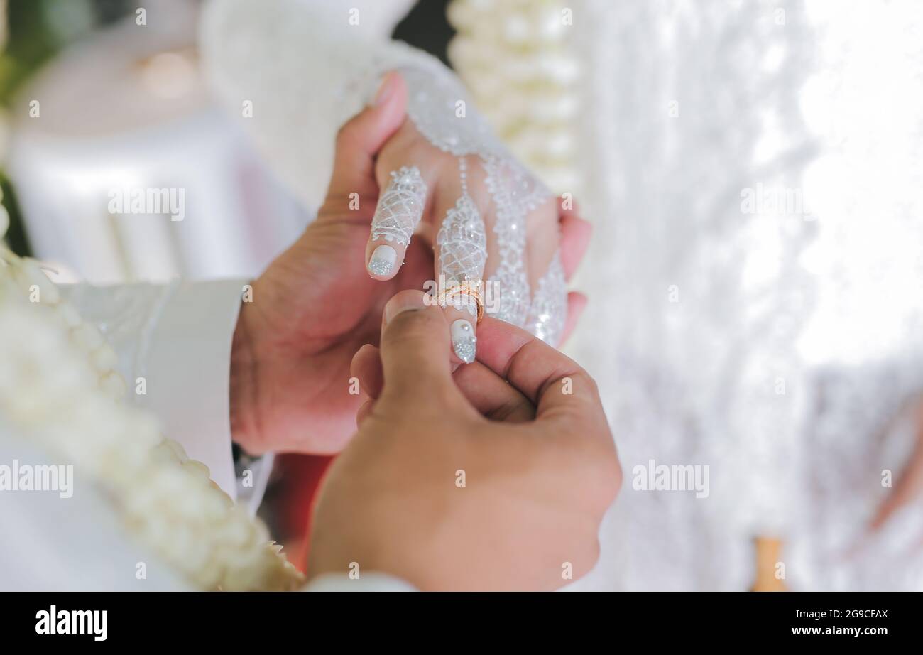 Ein flacher Fokus eines Bräutigams, der bei einer indonesischen Hochzeit einen Ehering auf die Hand der Braut mit weißem Henna-Tattoo legt Stockfoto