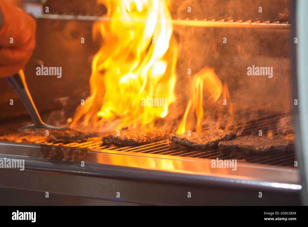 Flammen steigen durch Roste auf, während Hamburger zischen und auf dem Grill kochen. Stockfoto