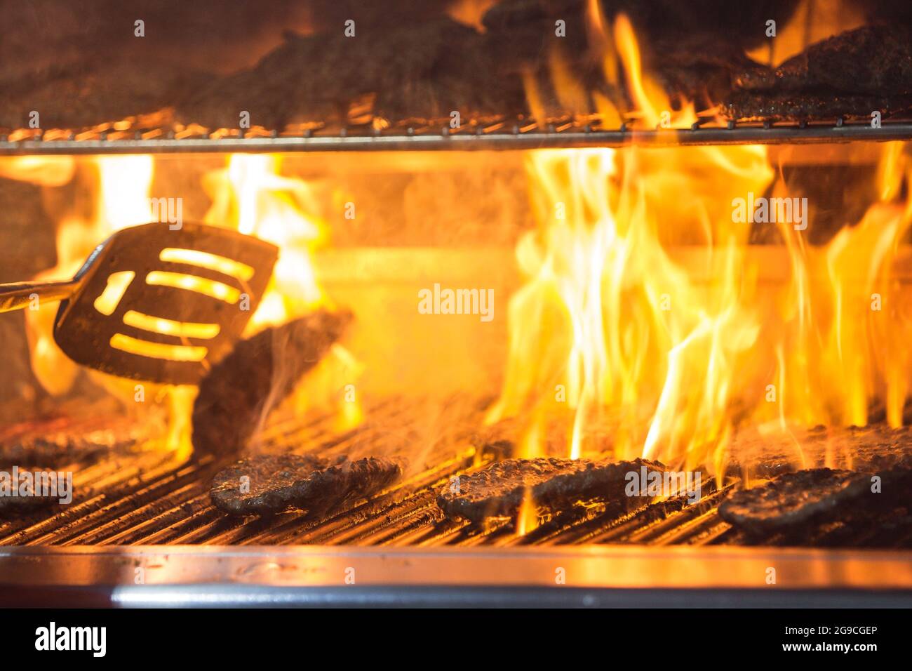 Goldene Flammen schießen durch die Grillroste, während Hamburger beim Kochen auf dem Grill zischen. Stockfoto