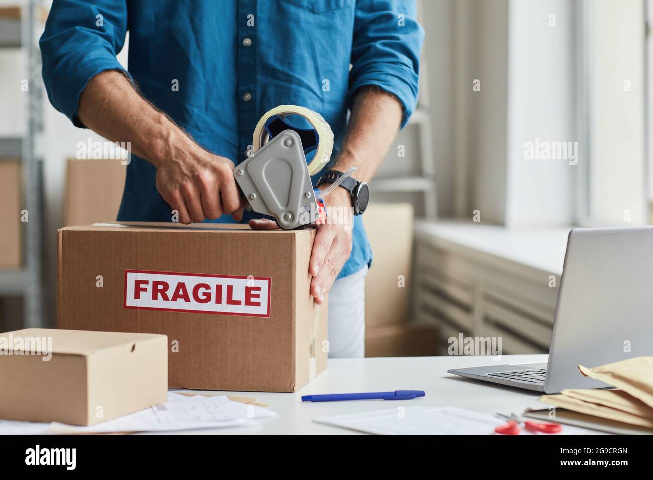 Ausgeschnittene Aufnahme von Mann, der im Lager mit einem Klebebandspender Kartons mit zerbrechlichen Aufklebern verpackt, Kopierfläche Stockfoto
