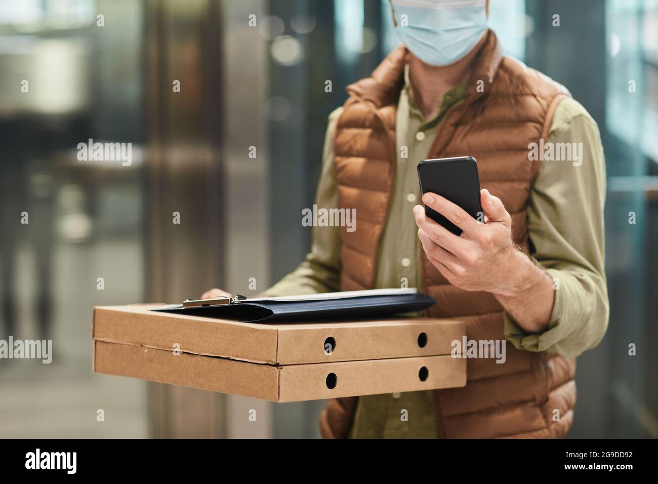 Beschnittenes Porträt eines Lieferers, der eine Maske trägt und Pizza hält, während er das Smartphone im Aufzug benutzt, Kopierraum Stockfoto