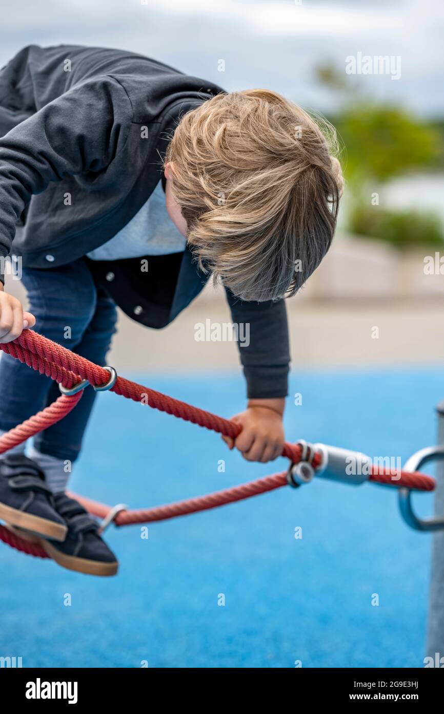 Der kleine Junge spielt begeistert mit einem gedehnten Seilnetz auf dem Spielplatz, um das Gleichgewicht des Kindes zu entwickeln und die motorischen Fähigkeiten zu entwickeln, die ihm die Möglichkeit geben Stockfoto