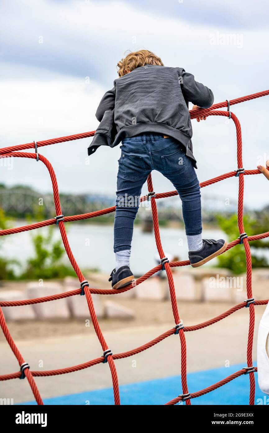 Der kleine Junge spielt begeistert mit einem gedehnten Seilnetz auf dem Spielplatz, um das Gleichgewicht des Kindes zu entwickeln und die motorischen Fähigkeiten zu entwickeln, die ihm die Möglichkeit geben Stockfoto