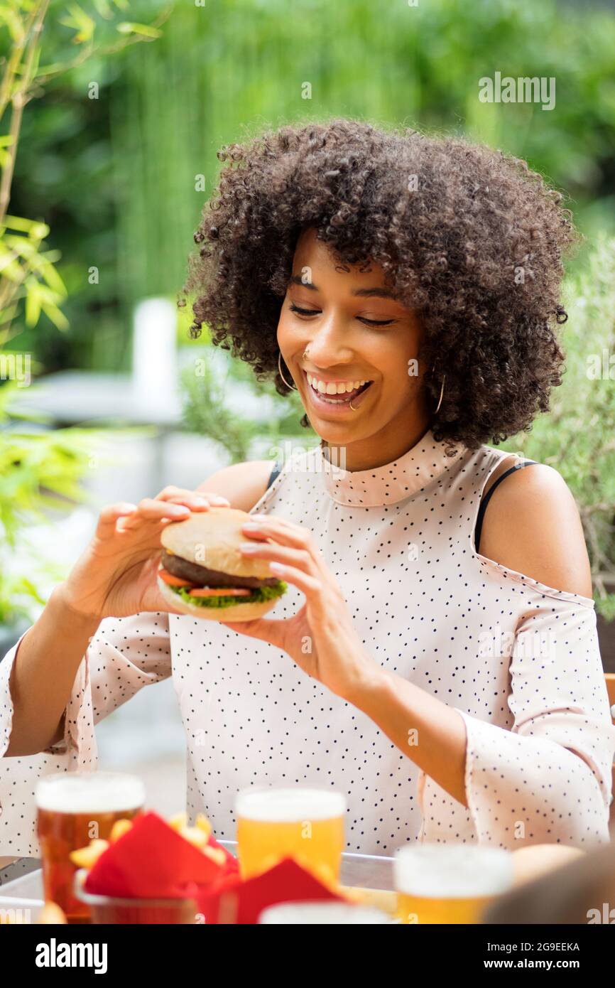 Lebhaft glückliche junge schwarze Frau, die einen Hamburger in ihren Händen mit einem strahlenden Lächeln und einem Blick der Vorfreude in einem Restaurant im Freien isst Stockfoto