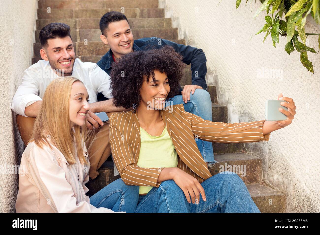 Eine Gruppe von glücklichen jungen Freunden, die auf einer Außentreppe für ein Selfie auf einem Mobiltelefon posieren, lachen und lächeln an der Kamera Stockfoto