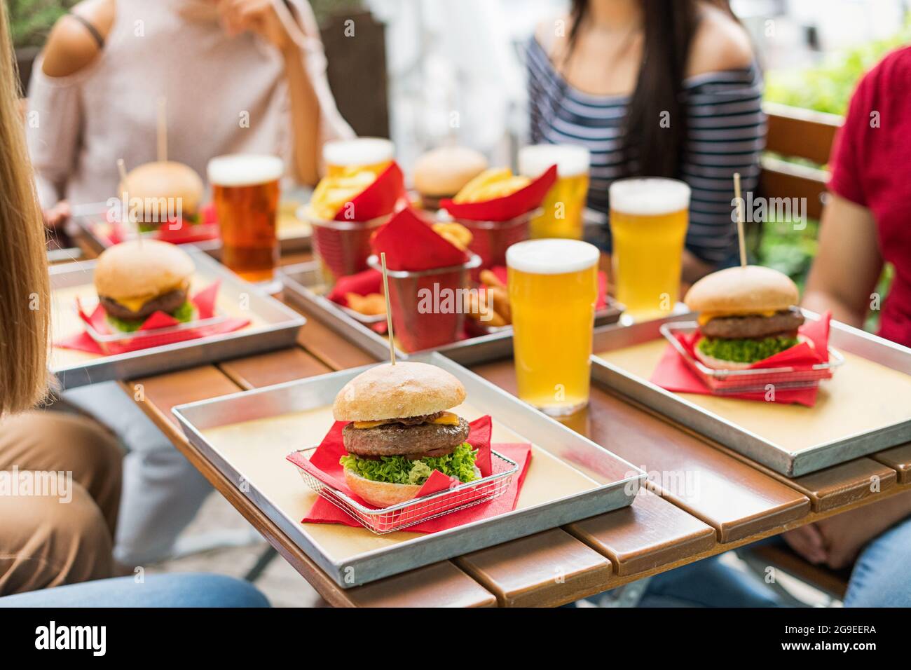 An einem Tisch im Freien werden Hamburger und kaltes Bier serviert, wobei anonyme Gäste um das Essen sitzen und sich gezielt auf einen Cheeseburger konzentrieren Stockfoto
