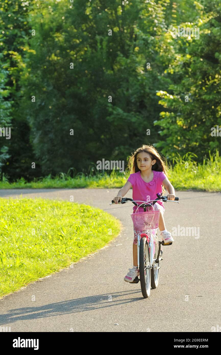 Ein junges Mädchen in einem stilvollen rosa Outfit, das mit dem Fahrrad auf einer kurvigen Landstraße im Frühlingssonne mit grünen Bäumen und Gras auf beiden Seiten fährt Stockfoto
