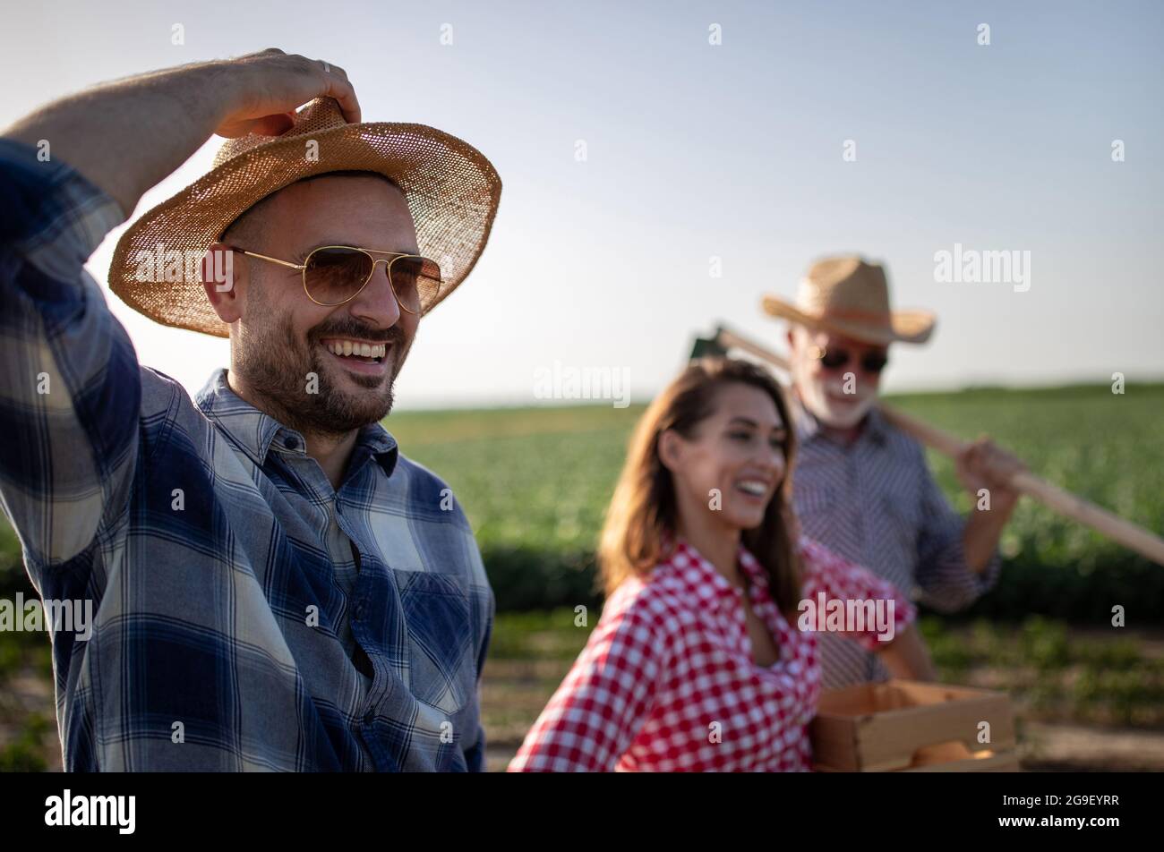 Junger Mann mit Sonnenbrille und lächelnder Hand auf dem Hut. Attraktive Bäuerin, die Kiste mit Produkten trägt. Älterer Mann mit Strohhut und Sonnenhut Stockfoto