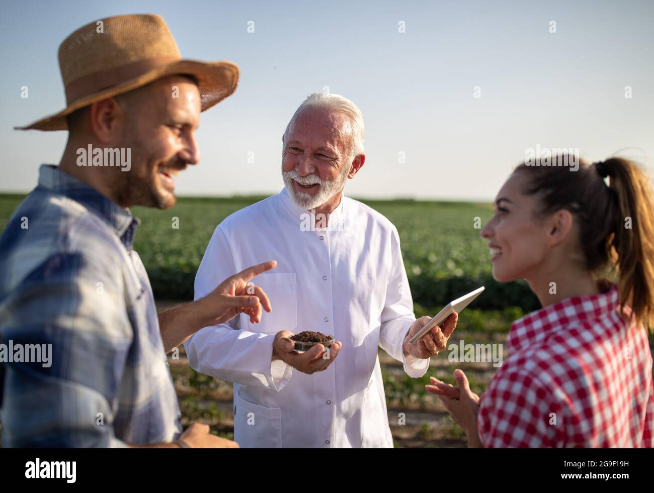 Paar junge Bauern diskutieren Bodenqualität stehen im Feld lächelnd. Ältere Wissenschaftler tragen Laborkittel mit Tablette und Petrischale mit Soi Stockfoto