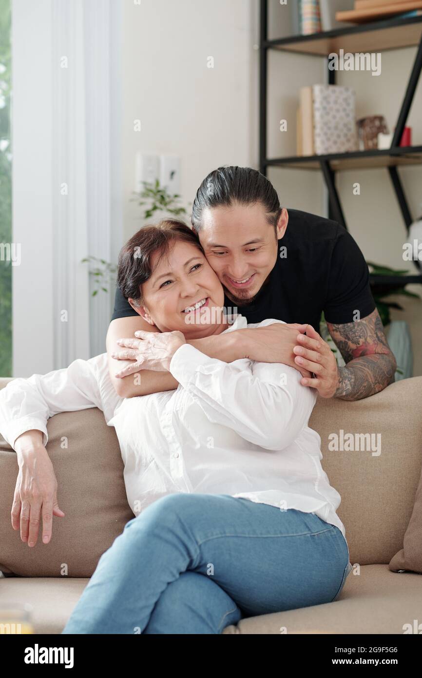 Älterer Sohn umarmt seine glückliche Mutter von hinten, während sie auf dem Sofa im Zimmer sitzt Stockfoto