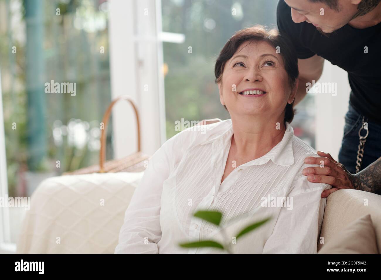 Asiatische Matue-Frau, die lächelnd auf dem Sofa sitzt und zu Hause mit ihrem älteren Sohn spricht Stockfoto