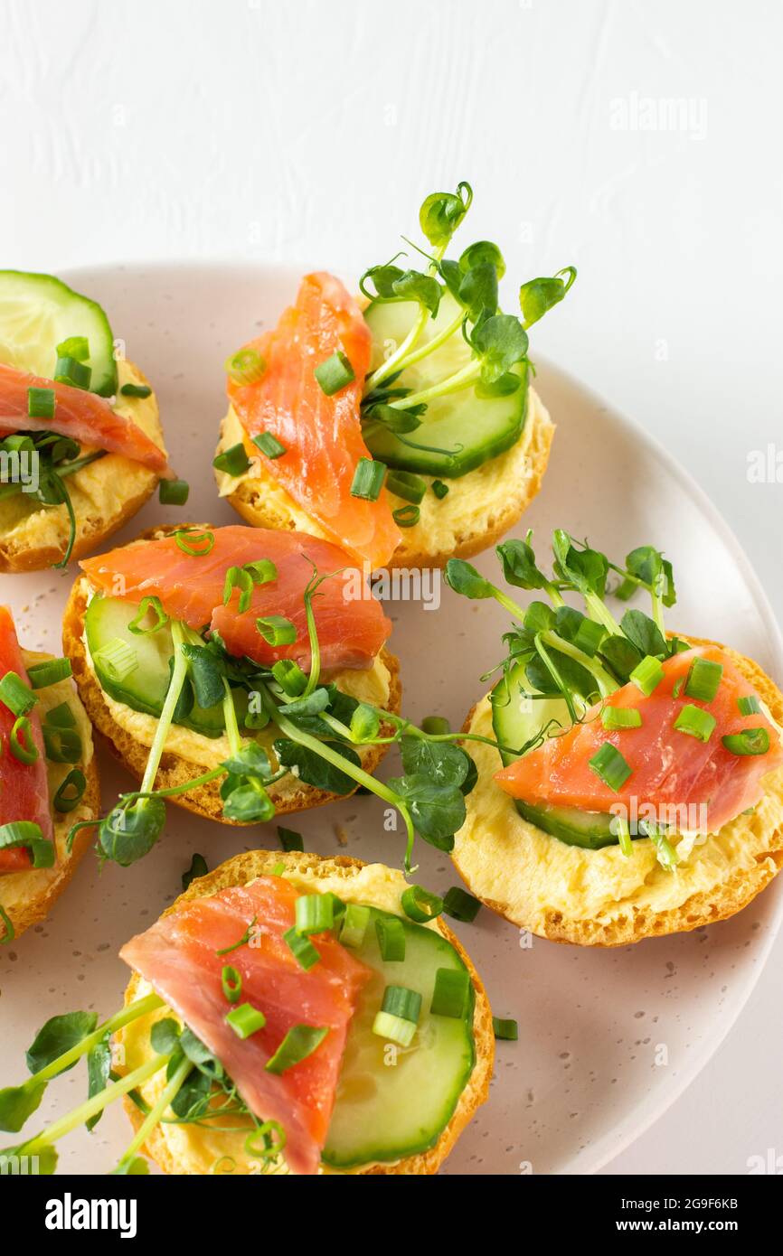 Leckere herzhafte Vorspeise, Lachsbraten auf einem weißen Teller, rundes gebratenes Brötchen mit rotem Fisch, Gurke, Sauce Hollandaise und Erbsensprossen Stockfoto