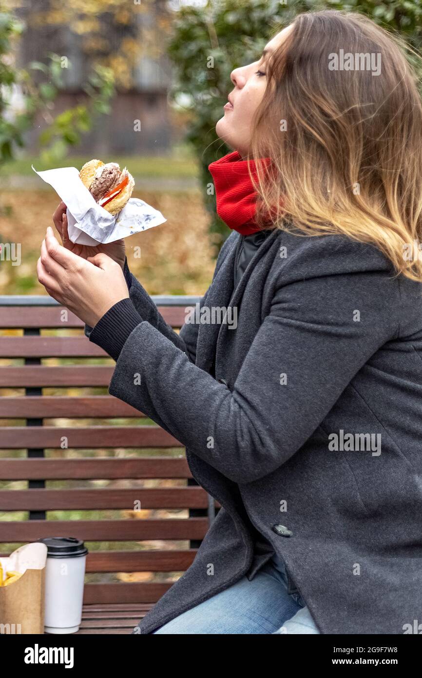 Junge hungrige Frau isst Burger und nimmt Mittagspause im Freien in Park.Fast Food. Essen zum Mitnehmen Konzept. Stockfoto