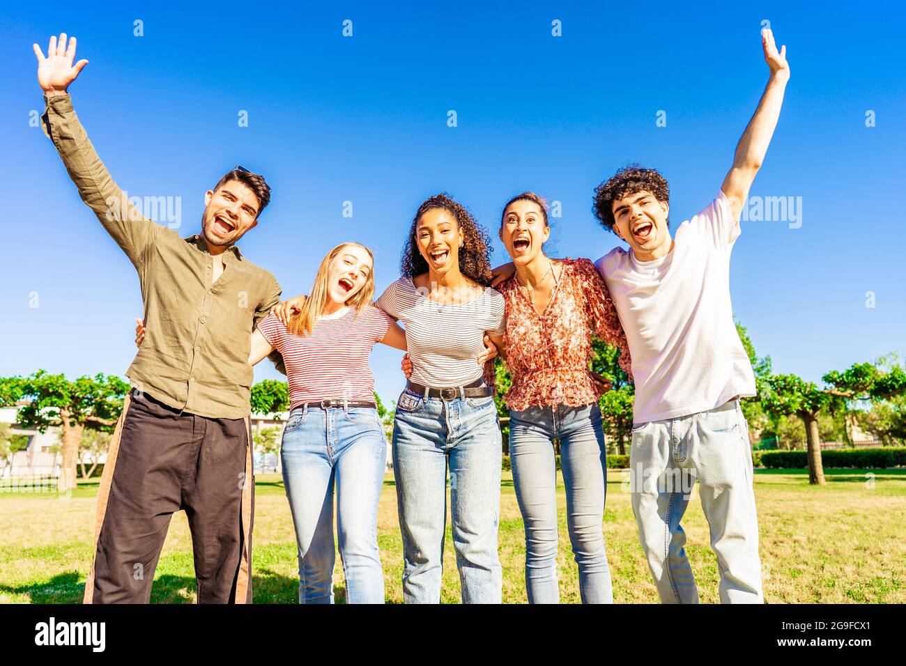 Multirassische Gruppe junger Freunde, die mit erhobenen offenen Armen posieren und die sich gegenseitig umarmende Kamera in der Natur des Stadtparks betrachten. Fröhliche, vielfältige Menschen Stockfoto