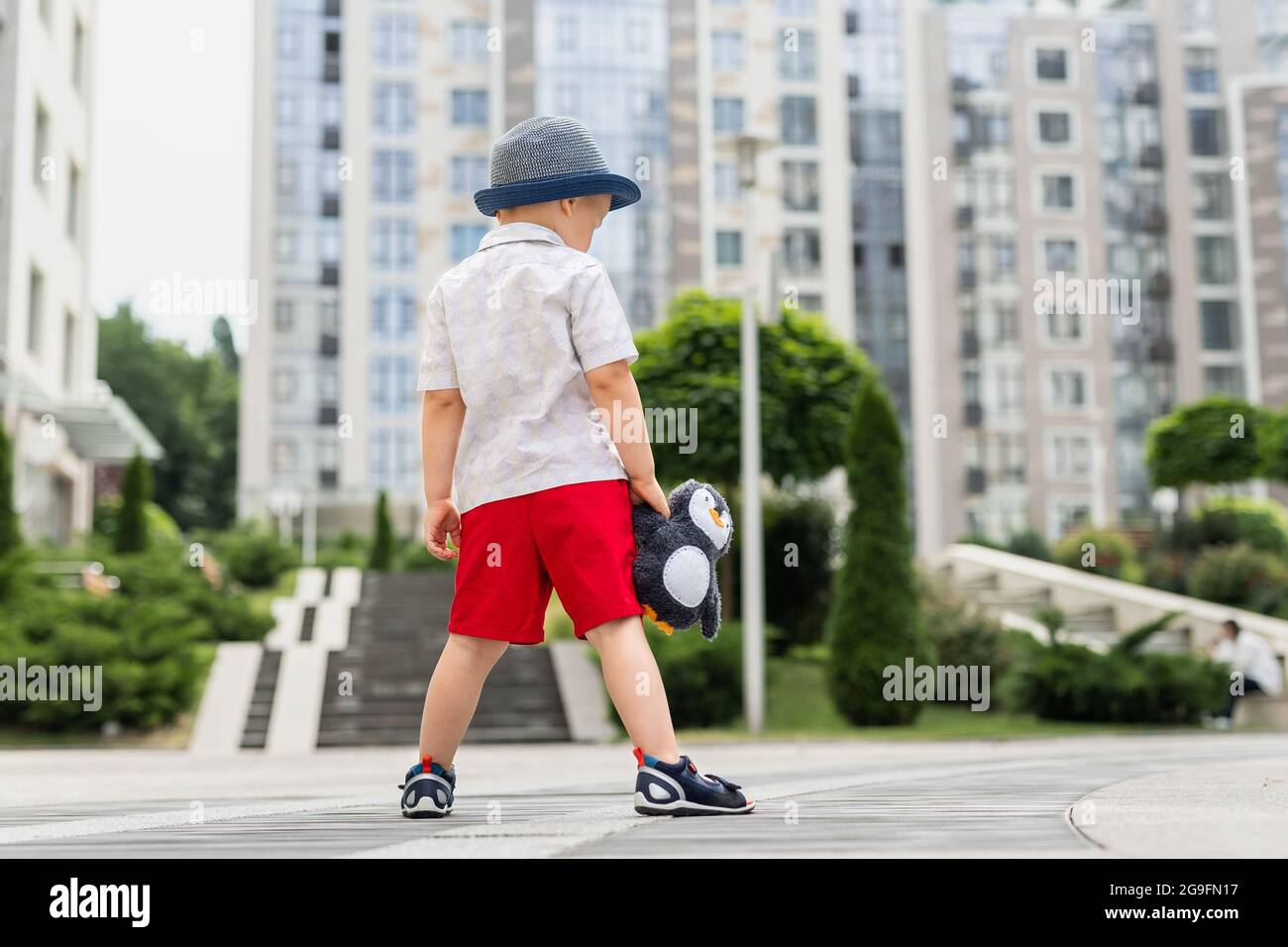 Rückansicht von niedlich allein traurig liebenswert kaukasischen 3-4 Jahre blonde kleine Junge in stilvollen Shorts Hemd und Hut halten weichen Teddy Spielzeug im Freien verloren Stockfoto