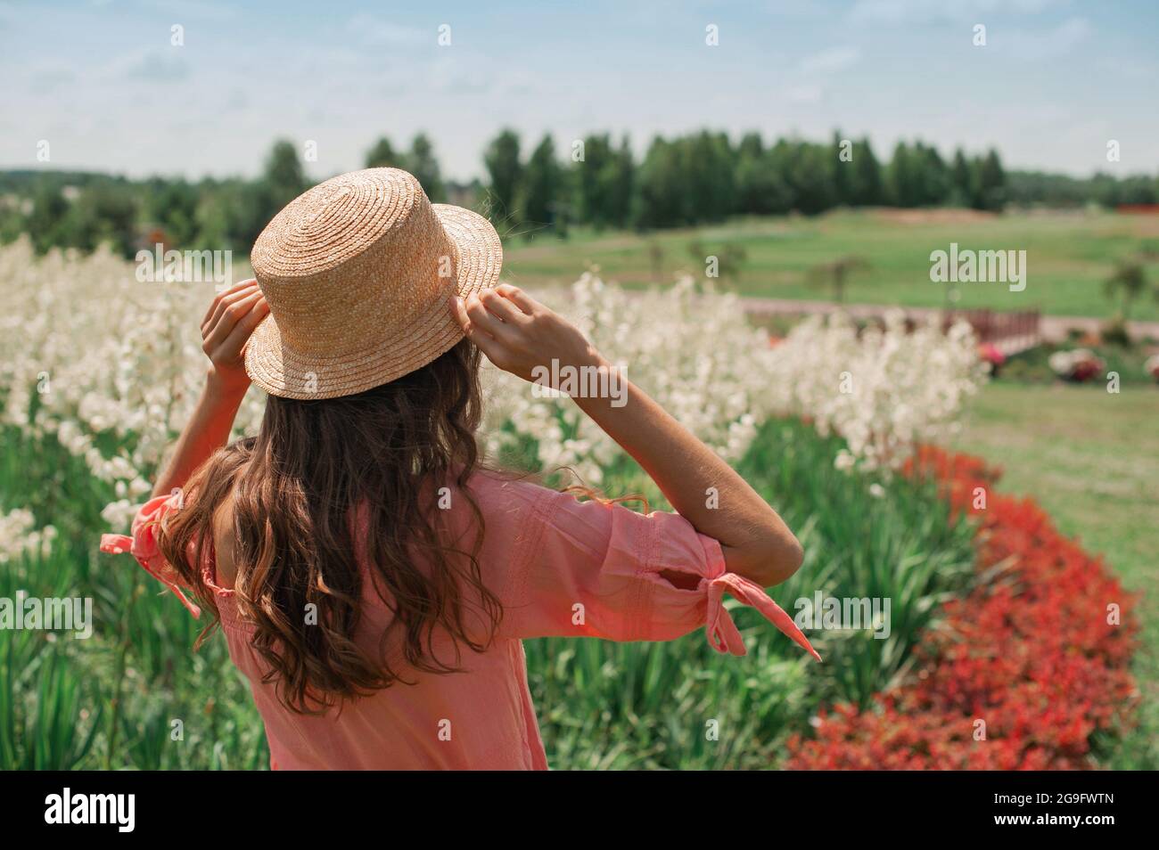 Frau in Hut in der Nähe von Blumenbeet. Mädchen in rosa Kleid, in Strohhut steht mit ihrem Rücken zur Sonne in floralen weißen und roten Feld, hält den Hut Stockfoto