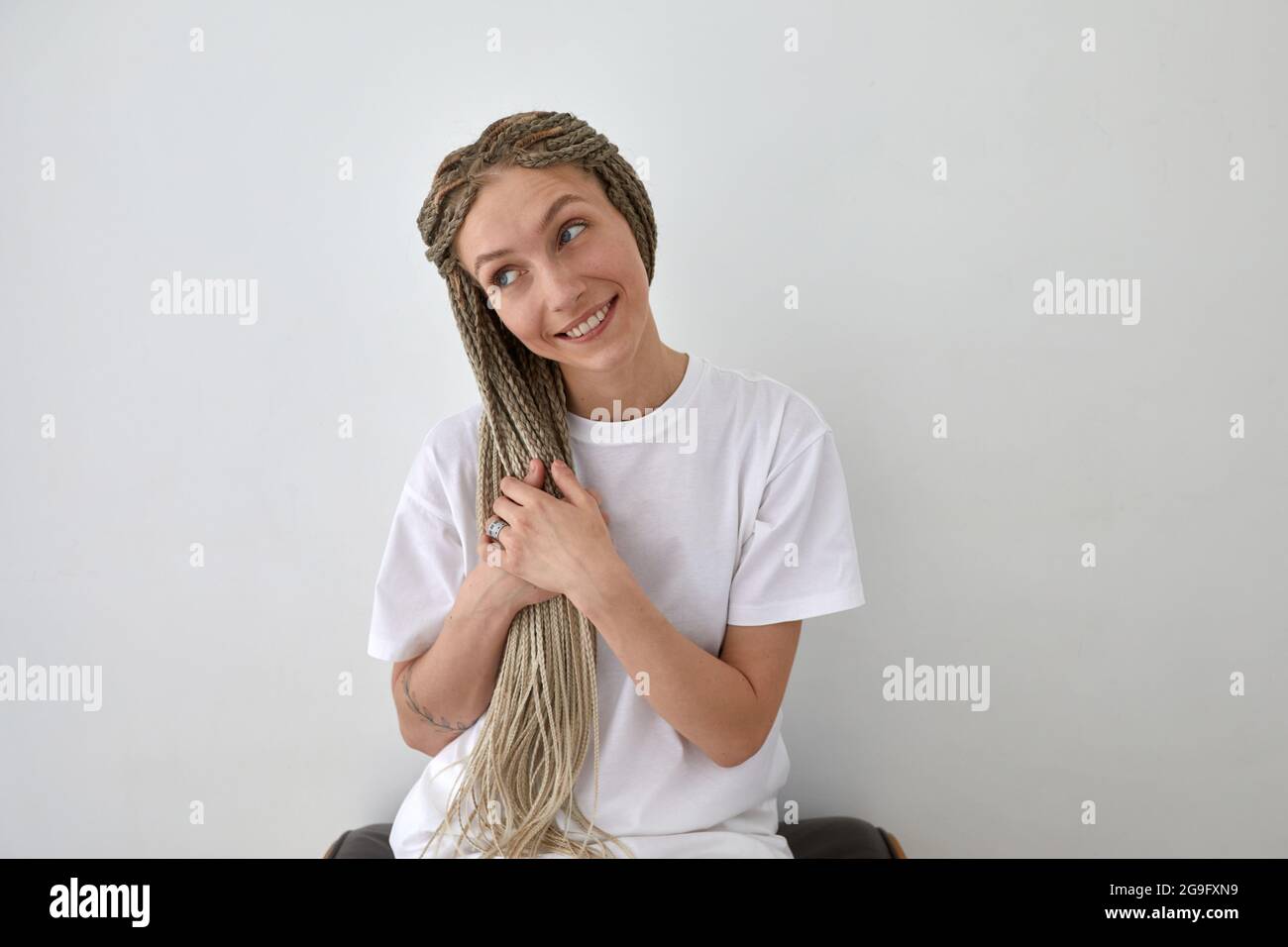 Positive Frau trägt ein weißes T-Shirt, das in der Nähe der Wand sitzt und Zöpfe berührt, während sie wegschaut Stockfoto