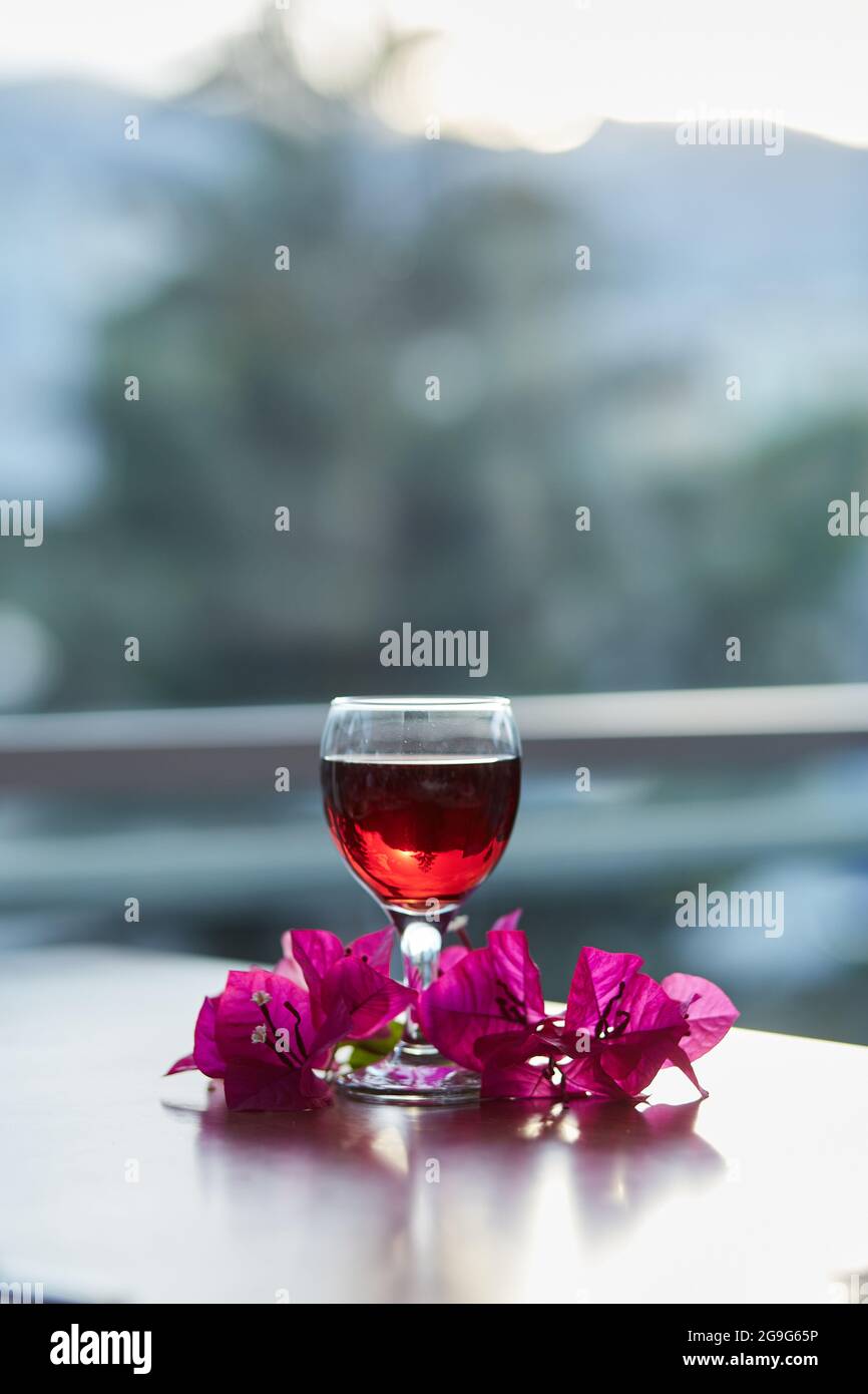 Ein Glas Rotwein auf unscharfem Berghintergrund. Dekorationen von rosa Bougainvillea Blumen. Blue Hour Fotografie. Speicherplatz kopieren. Stockfoto