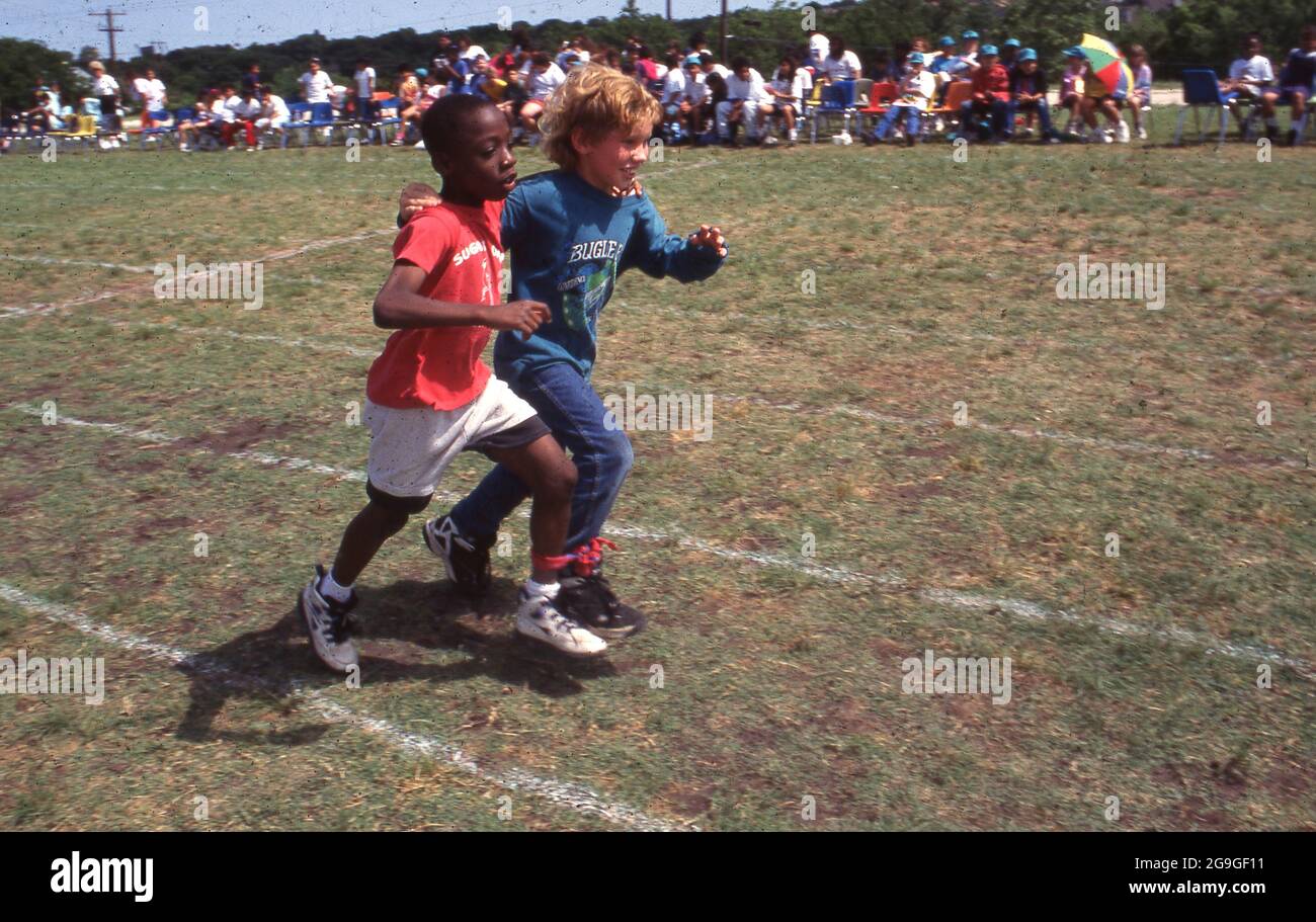 Austin, Texas, USA, 1990: Schwarzer Junge und weiße Teamkollegen, deren Knöchel zusammengebunden sind, laufen während des Feldtages an ihrer Grundschule im dreibeinigen Rennen. ©Bob Daemmrich Stockfoto