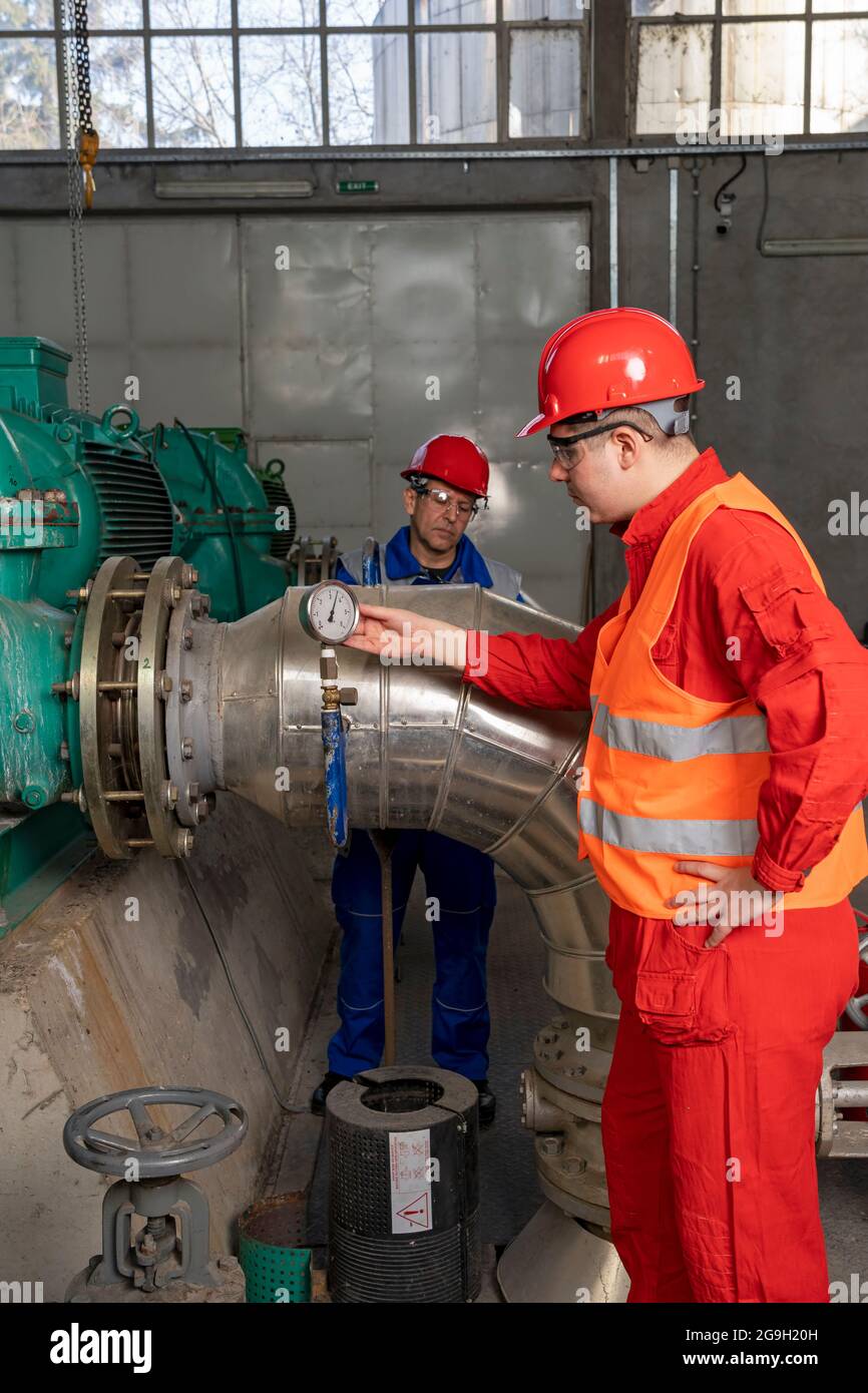 Junge Arbeiter in persönlichen Schutzausrüstung Überprüfung Manometer Instrument.zwei Arbeiter stehen neben großen Generatoren in industriellen Innenraum. Stockfoto