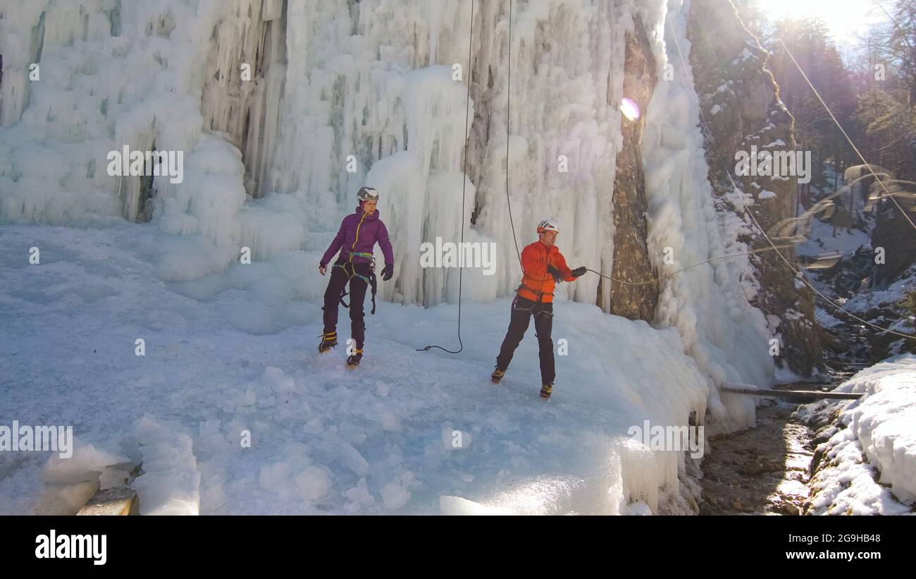 Handgeführte Aufnahme eines kaukasischen Paares, Eiskletterer in Schutzkleidung, die am Fuß des gefrorenen Wasserfalls stehen und sich auf den nächsten Aufstieg vorbereiten Stockfoto