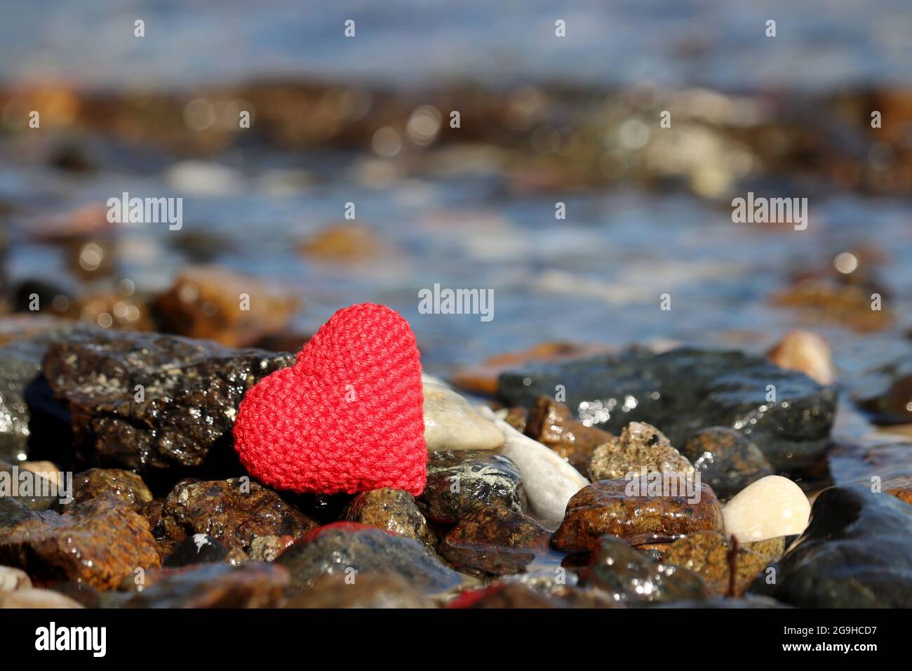 Liebe am Strand, rot gestricktes Herz auf einem Kieselstein auf Meeresgrund. Konzept von Valentinstag, romantischem Sommer oder Flitterwochen Stockfoto