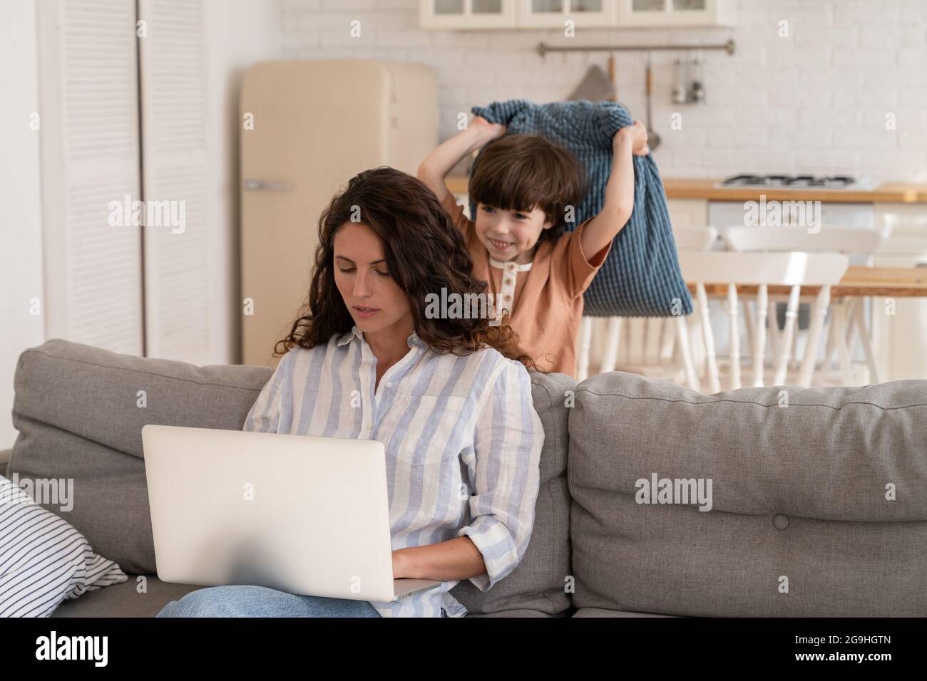 Arbeiten Sie von zu Hause aus mit einem frechen Kind: Junge Mama nutzt einen Laptop für geschäftliche Zwecke Sitzen Sie auf dem Sofa mit einem gelangweilten Sohn-Angriff Stockfoto