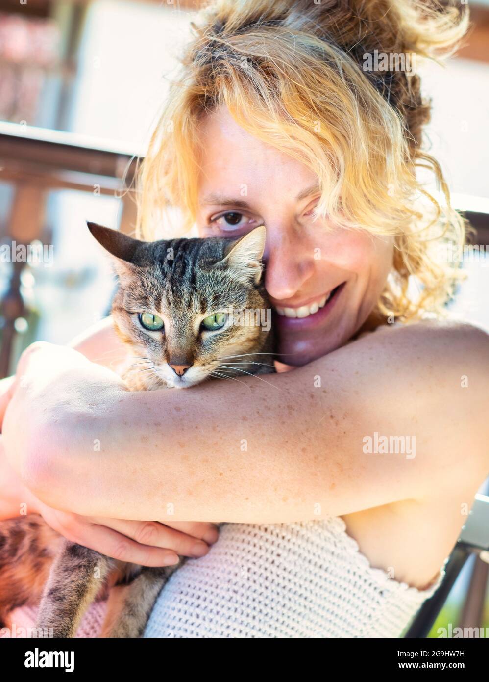 Porträt einer jungen reifen kaukasischen blonden Frau, die mit ihrer Katze zu Hause auf der Veranda lächelt und in einem Schaukelstuhl sitzt. Lifestyle-Konzept. Stockfoto