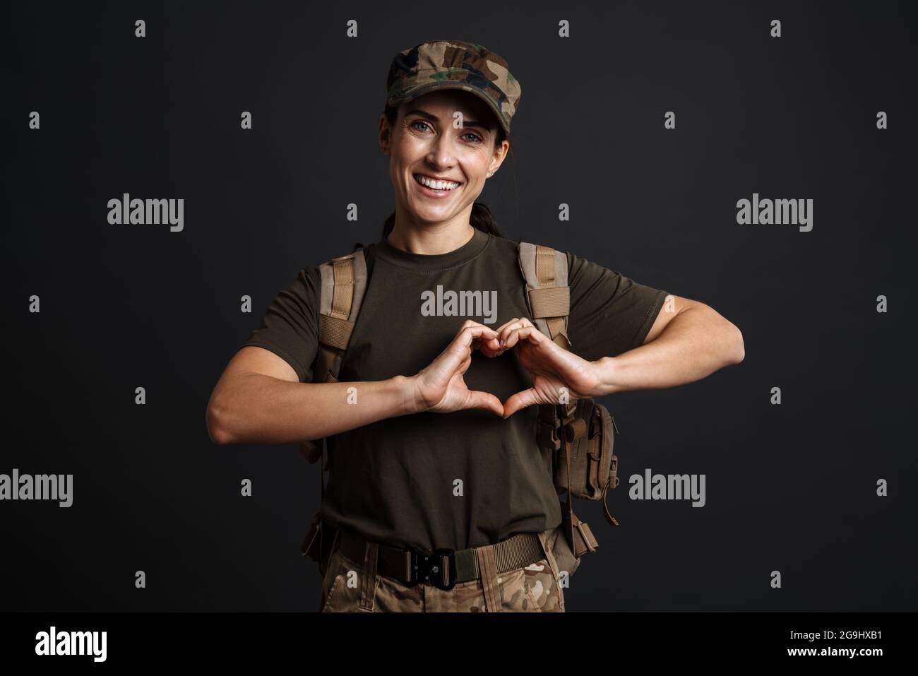 Lächelnde Soldatin mit Armeerucksack, die eine vereinzelte Herzbewegung auf schwarzem Hintergrund zeigt Stockfoto
