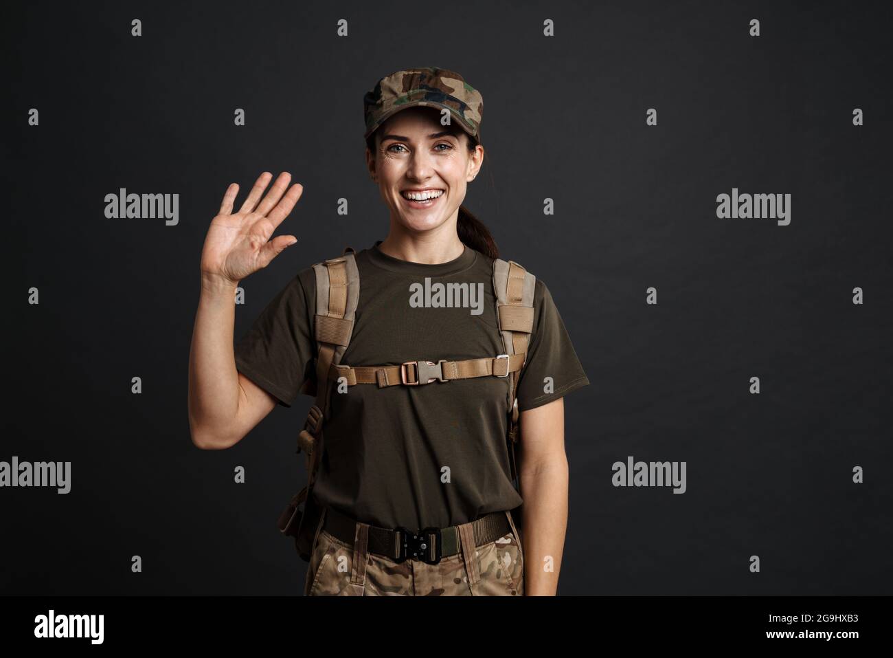 Lächelnd schöne Soldatin mit Armee Rucksack winkende Hand isoliert auf schwarzem Hintergrund Stockfoto