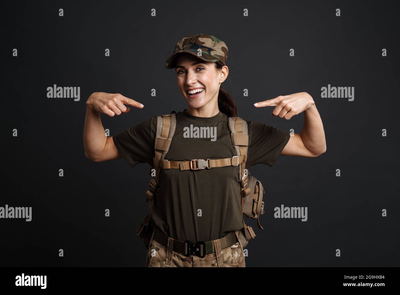 Lächelnde Soldatin mit Armeerucksack, der die Finger auf sich selbst zeigt, isoliert auf schwarzem Hintergrund Stockfoto