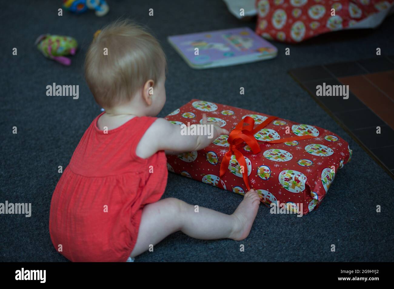 Niedliches Baby Mädchen mit roten Kleidern sitzt auf dem Boden und öffnet eine Geschenk-Box. Stockfoto