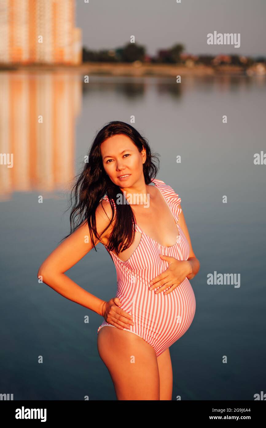 Schwangere in einem Badeanzug, entspannte, dunkelhaarige Schwangere in  einem rosa Badeanzug hält ihre Hände auf ihrem schwanger Bauch in der Bucht  bei Sonnenuntergang auf einem Stockfotografie - Alamy