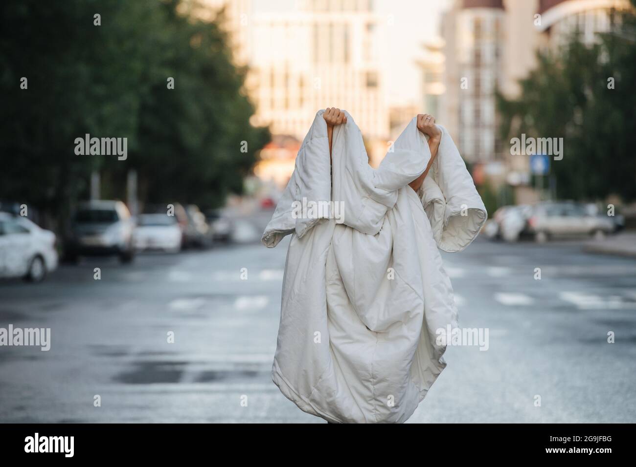 Person in Decke in der Mitte der leeren nassen Stadtstraße am frühen Morgen gewickelt. Ghost wird gespielt. Stockfoto