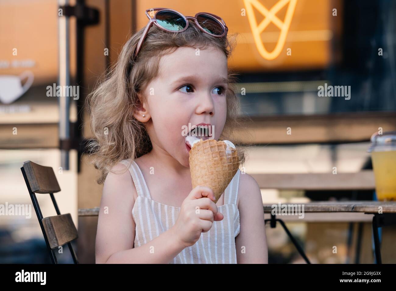 Kleinkind, das draußen in der Nähe des Cafés Kegeleis isst. Cute blonde kaukasische Mädchen lecken gefrorenen Sommer Essen auf der Straße. Schmutziger Mund, glückliches Kind. Stockfoto