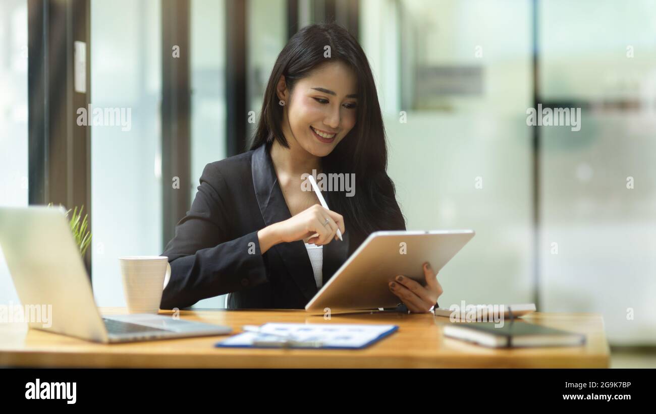 Schöne elegante Geschäftsfrau in schwarzem Anzug lächeln, während Blick auf Finanzbericht über Tablet und halten Stift, Laptop, Rechner und Dokument auf Holz de Stockfoto