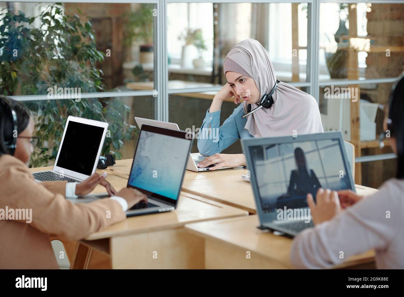 Müde muslimische Geschäftsfrau, die vor dem Laptop unter ihren Kollegen am Schreibtisch sitzt Stockfoto