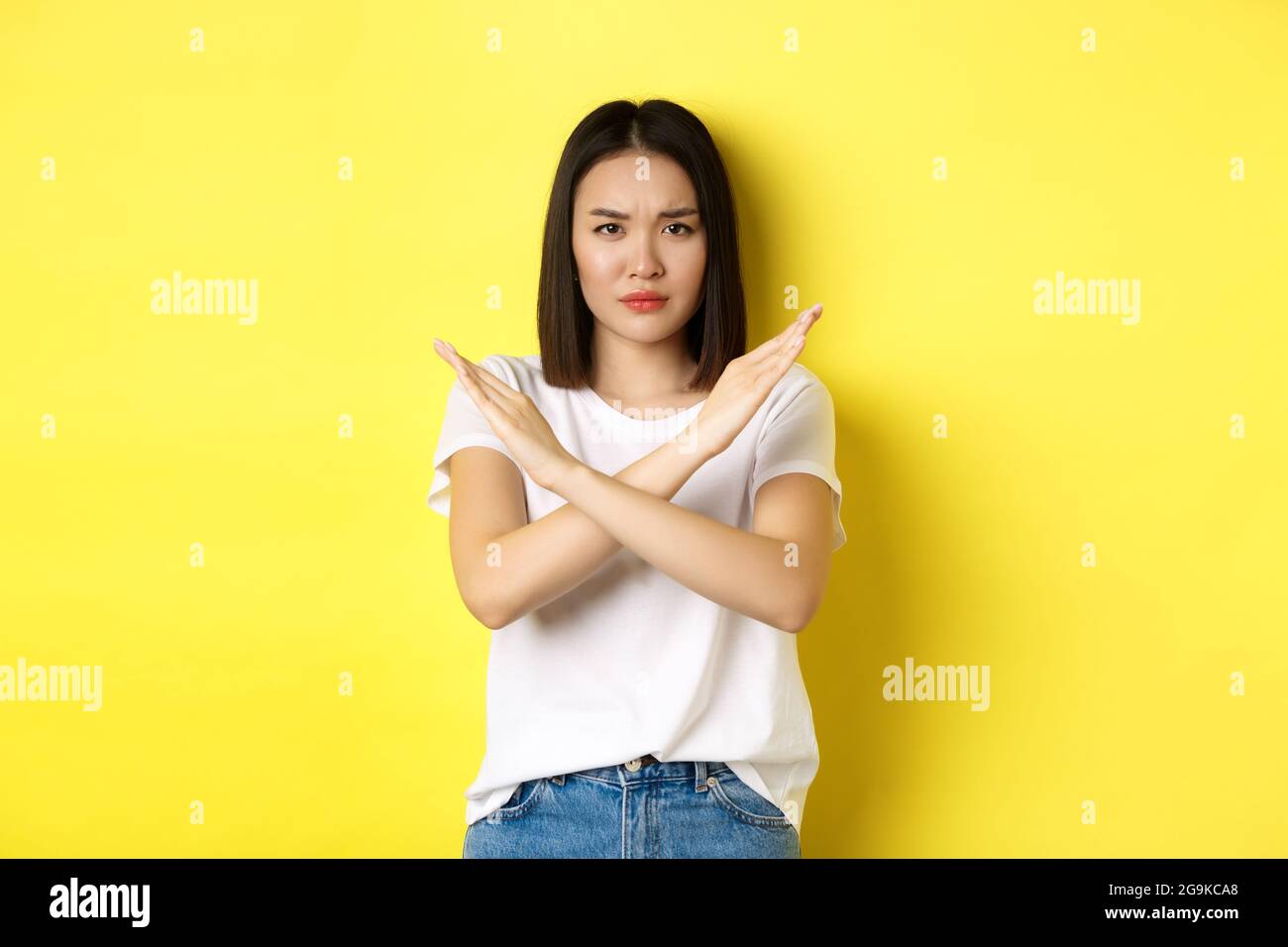 Seriöse und durchsetzungsfähige koreanische Frau mit Kreuzstoppgeste, Stirnrunzeln und Nein sagen, Aktion verbieten, etwas Schlechtes missbilligen, über Yello stehen Stockfoto