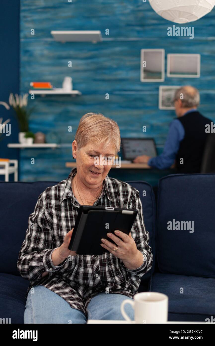 Rentnerin im Internet sitzend auf der Couch. Ältere Frau mit Modder Technoloy Tablet-pc im Wohnzimmer zu Hause und Mann arbeiten an Laptop-Computer Stockfoto