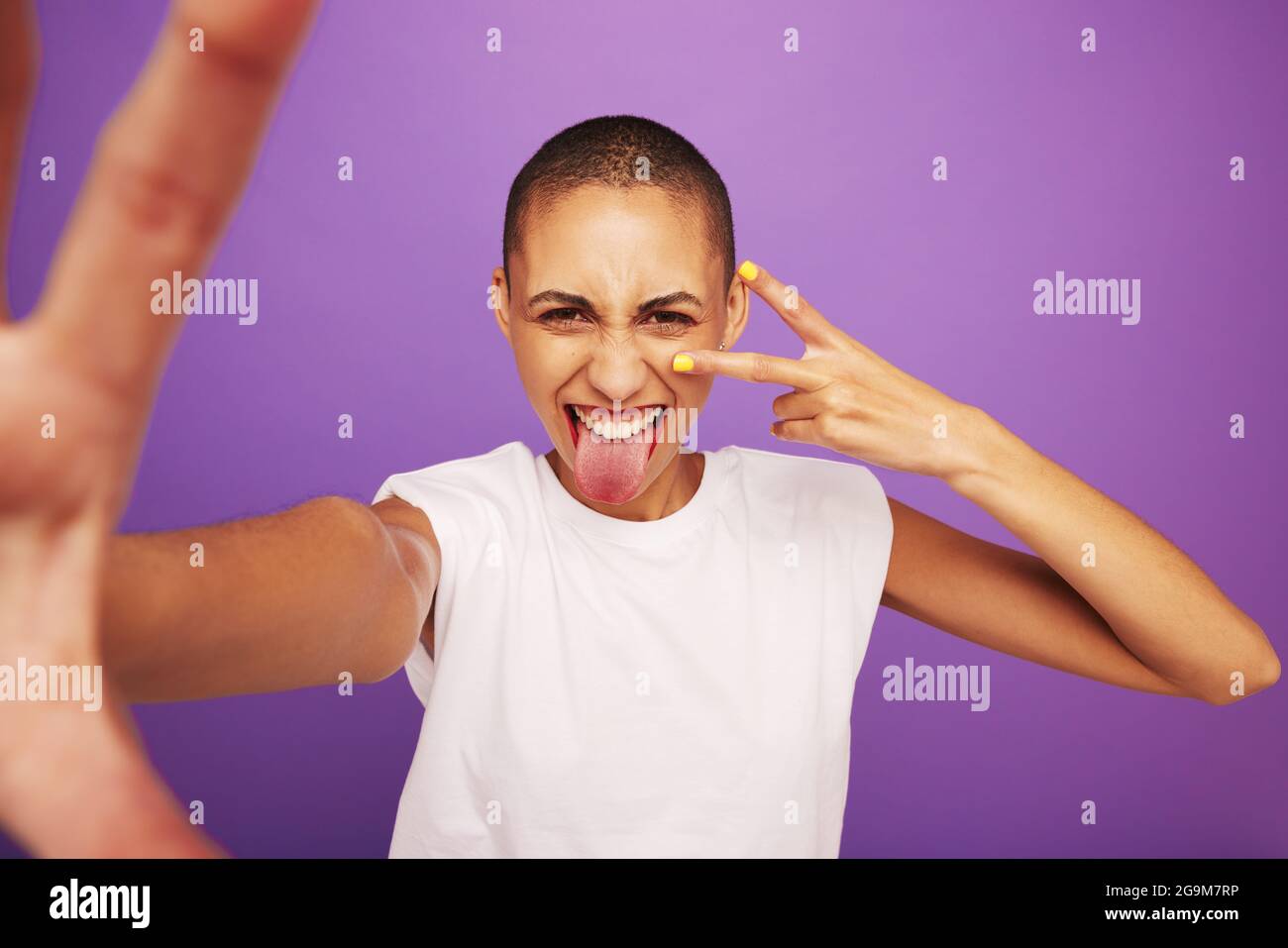 Porträt einer ausdrucksstarken Frau, die auf violettem Hintergrund posiert. Rasierte Kopffrau, die ihre Zunge herausstreckt und ein Zeichen der Friedenshand zeigt. Stockfoto