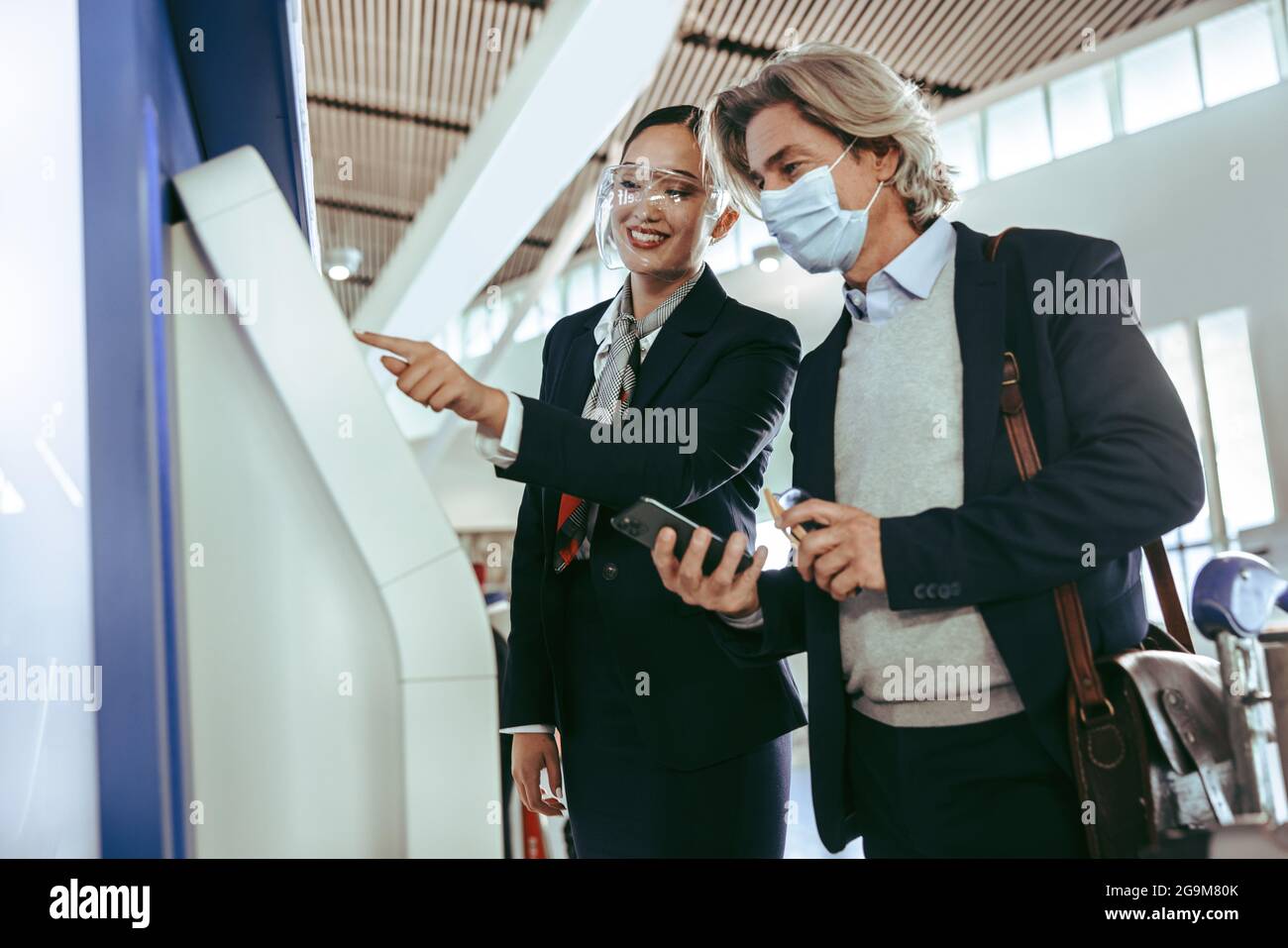 Eine weibliche Bodenbegleiterin mit Gesichtsschutz hilft männlichen Reisenden mit einem Check-in-Automaten am Flughafenterminal während einer Pandemie. Stockfoto